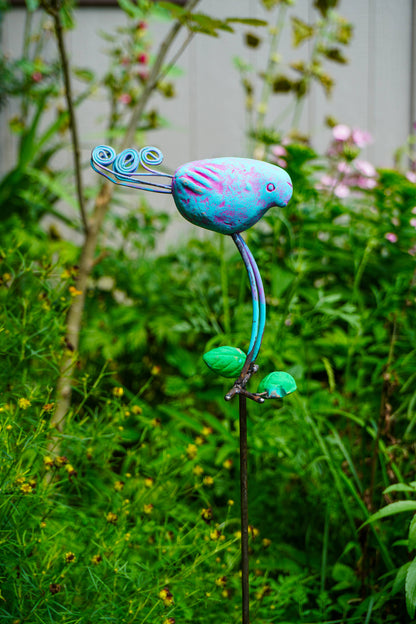 A colorful metal garden sculpture of a stylized bird perched on a stem with leaves, photographed outdoors in a garden setting with green foliage and blurred plants in the background.