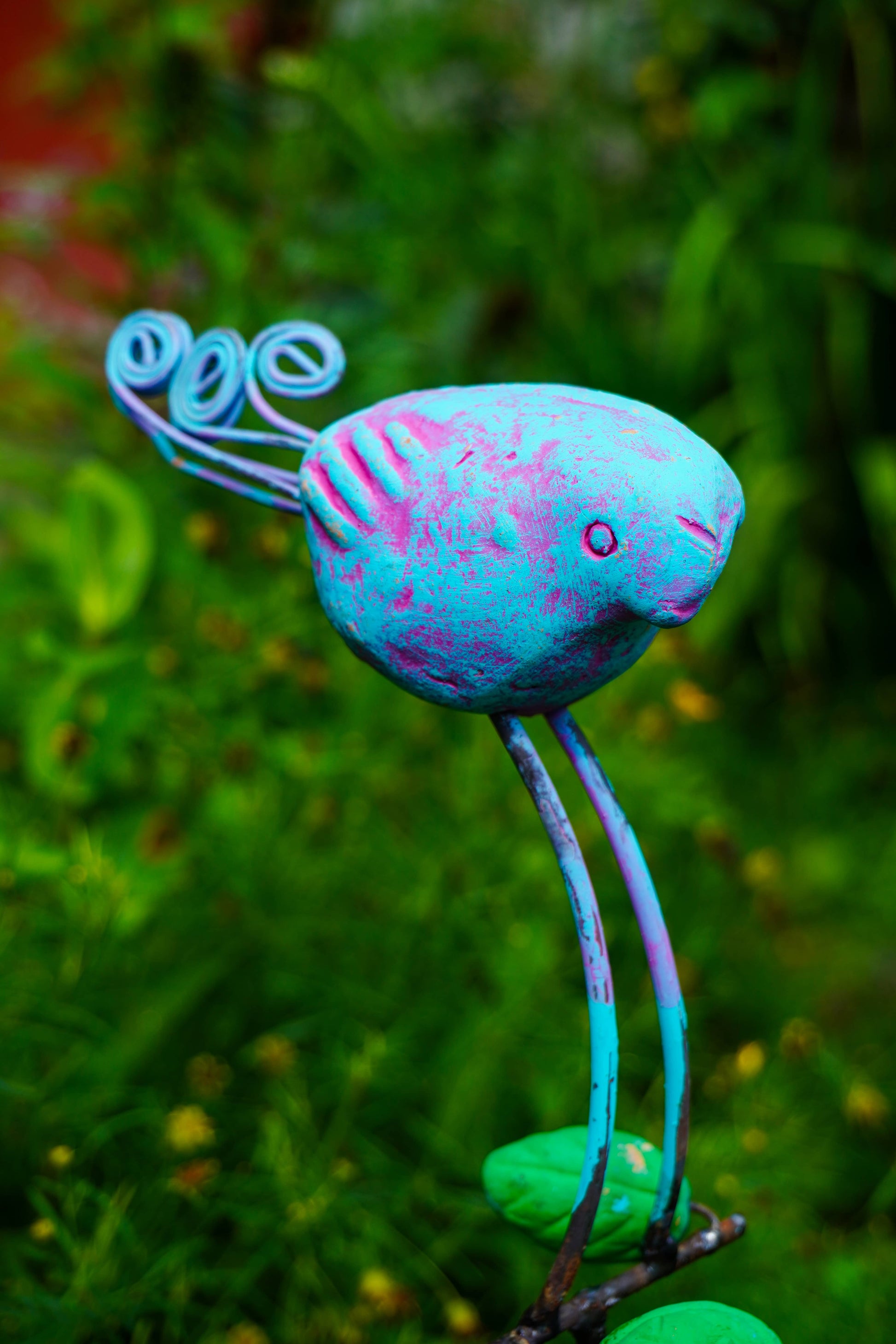 A colorful metal garden stake sculpture shaped like a stylized bird perched on a curved stem, photographed outdoors in a grassy garden path.