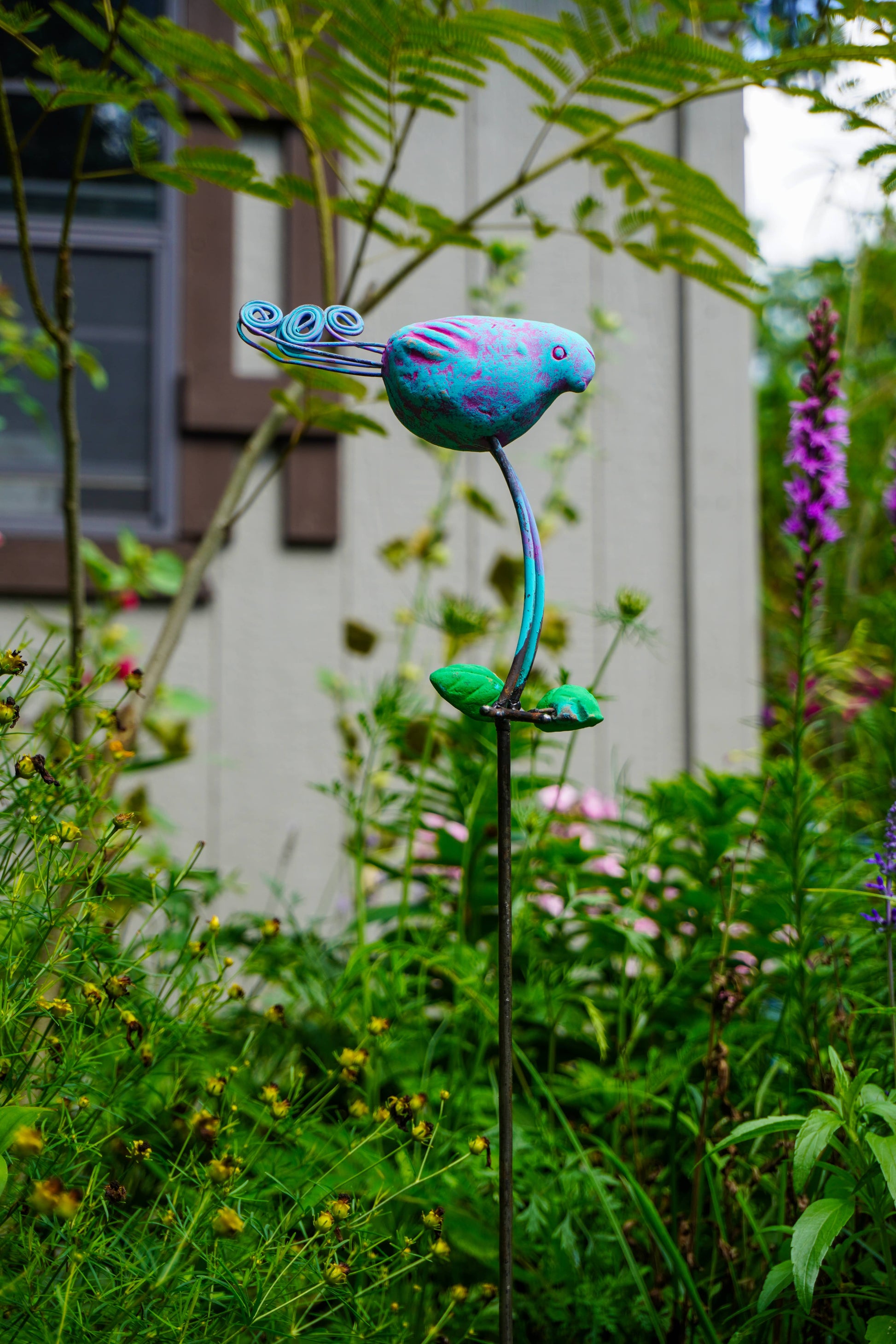A colorful metal garden stake sculpture shaped like a stylized bird perched on a curved stem, photographed outdoors in a grassy garden path.