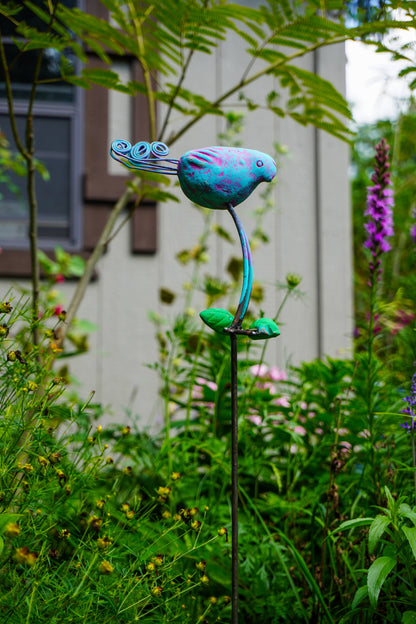 A colorful metal garden stake sculpture shaped like a stylized bird perched on a curved stem, photographed outdoors in a grassy garden path.