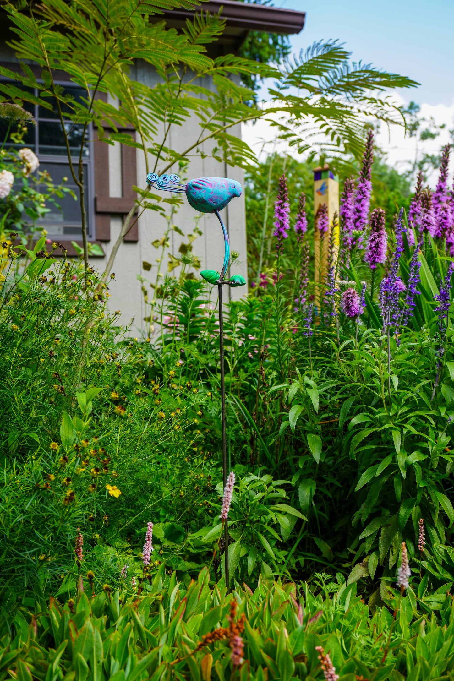 A garden scene with dense green foliage, purple flowers, and a blue bird-shaped metal garden stake in the center.