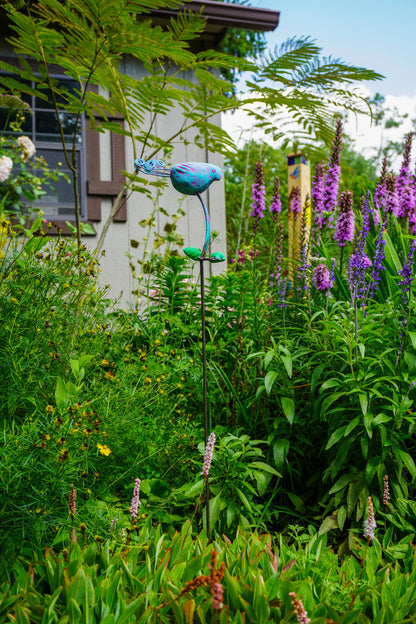 A garden scene with dense green foliage, purple flowers, and a blue bird-shaped metal garden stake in the center.