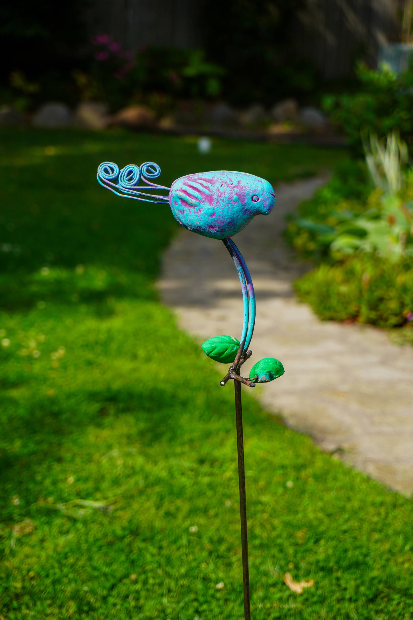 A colorful metal garden stake sculpture shaped like a stylized bird perched on a curved stem, photographed outdoors in a grassy garden path.