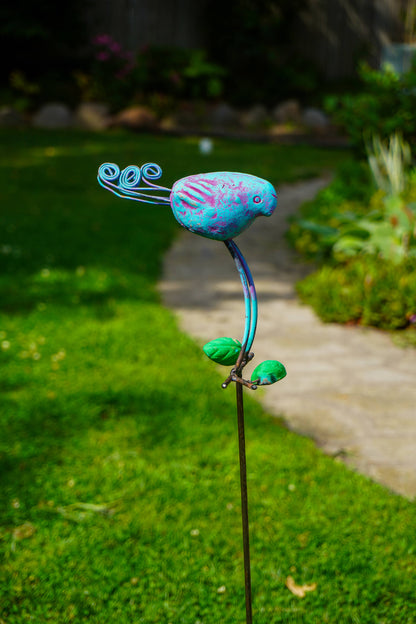 A colorful metal garden stake sculpture shaped like a stylized bird perched on a curved stem, photographed outdoors in a grassy garden path.
