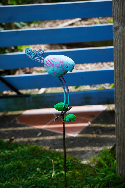A colorful metal garden stake sculpture shaped like a stylized bird perched on a curved stem, photographed outdoors in a grassy garden path.