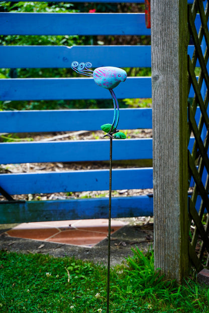 A colorful metal bird garden stake sculpture with a blue and pink flower head, green leaves, and a long thin stem, photographed outdoors in front of a blue wooden fence and wooden lattice.