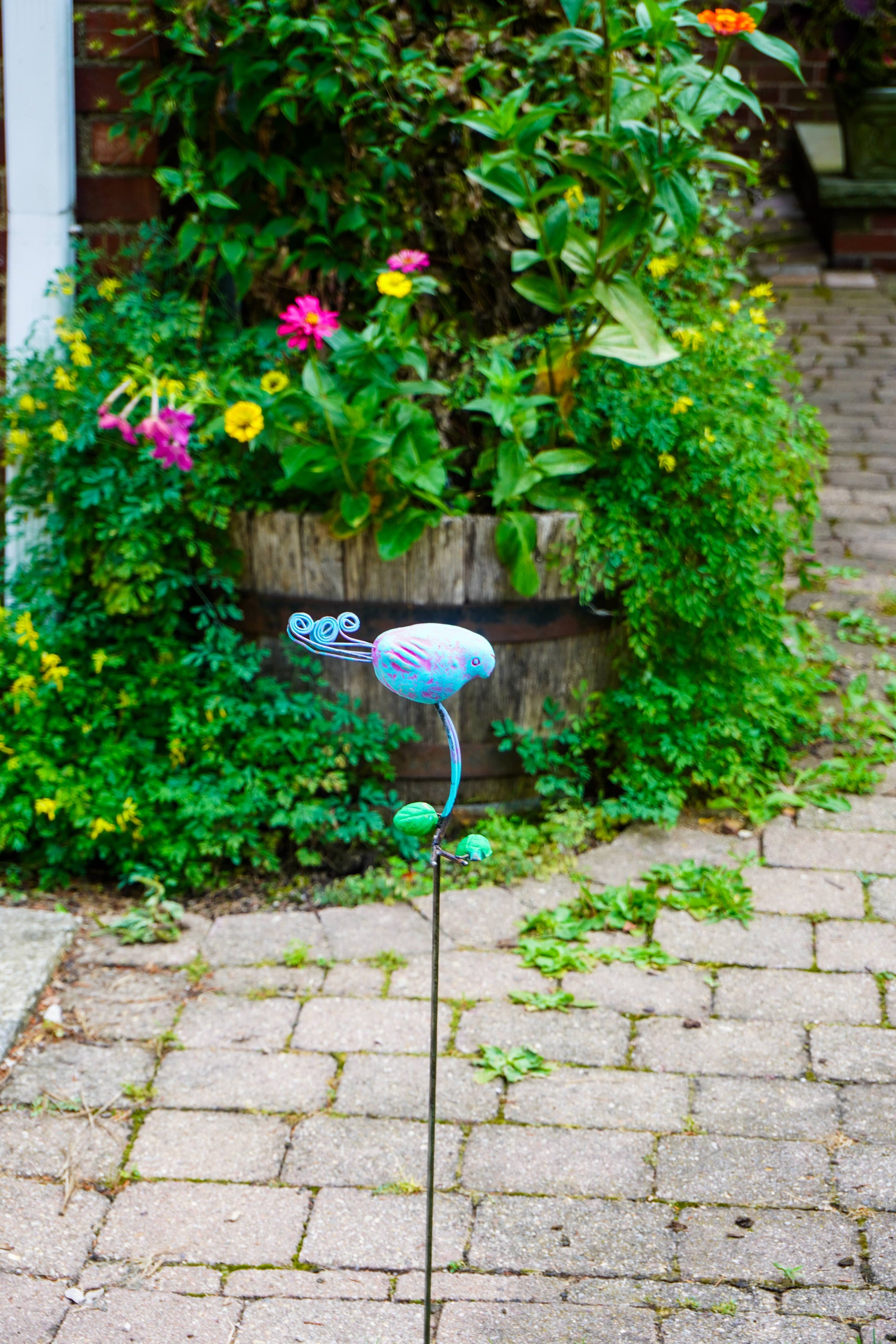 A colorful metal garden stake sculpture shaped like a bird, placed on a paved path in front of a lush green garden bed with flowers.