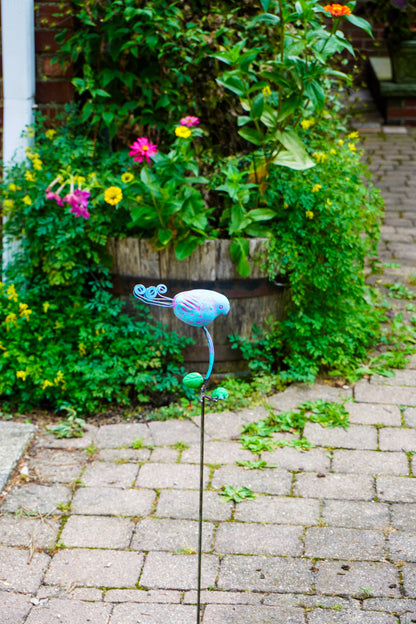 A colorful metal garden stake sculpture shaped like a bird, placed on a paved path in front of a lush green garden bed with flowers.