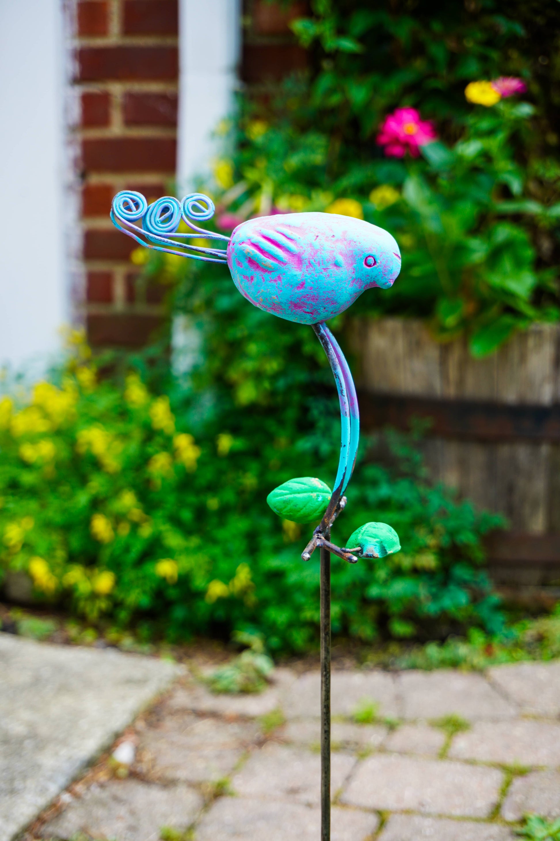 A colorful metal garden stake sculpture shaped like a stylized bird perched on a curved stem, photographed outdoors in a grassy garden path.