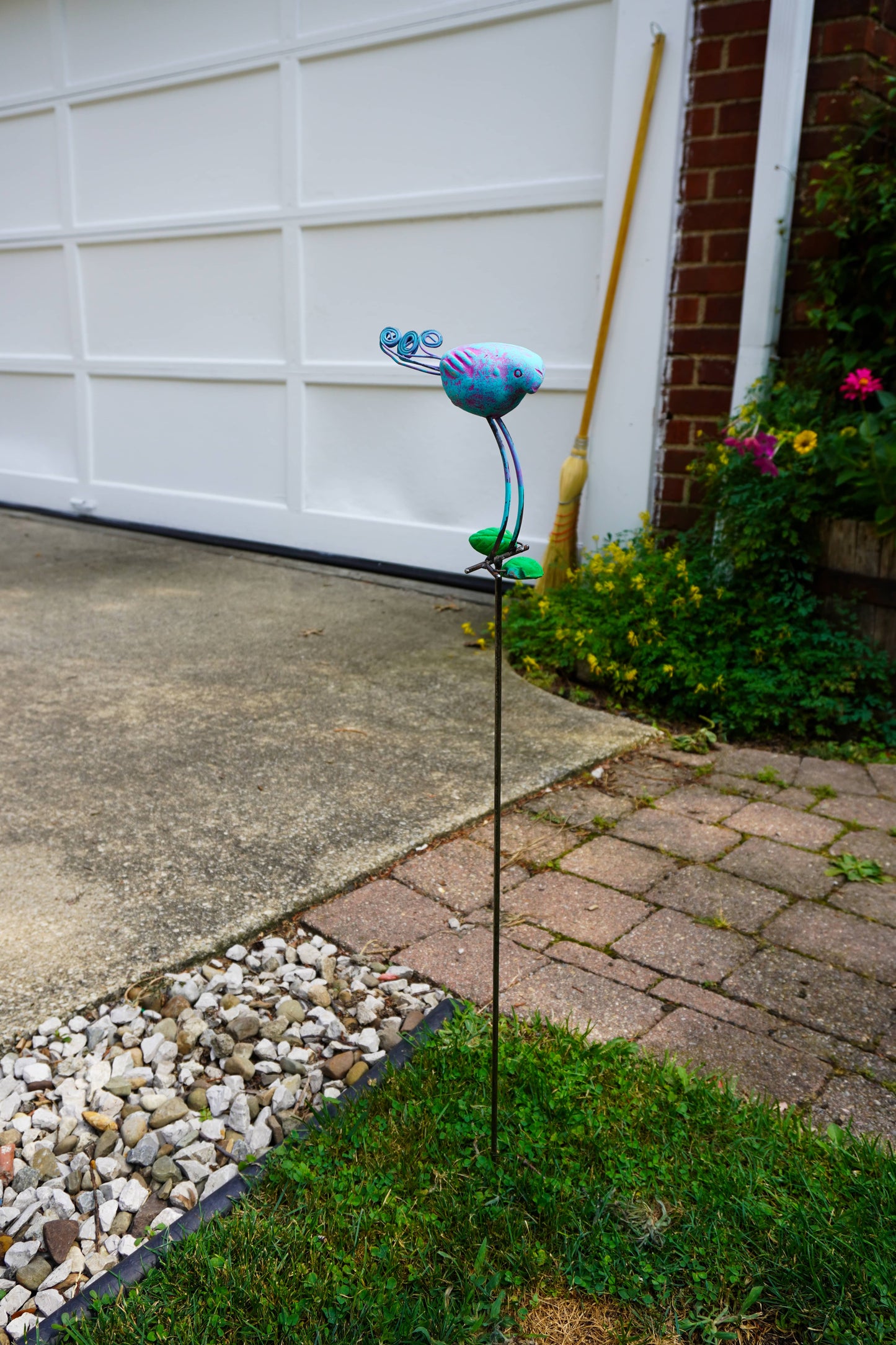 A colorful metal garden stake sculpture shaped like a stylized bird or flower, placed on grass in a backyard next to a white garage door, brick pavers, and a flower bed.