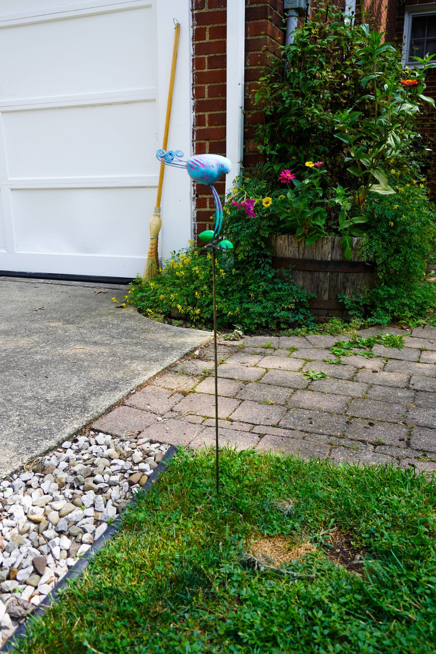 A metal garden stake with a colorful bird ornament stands in a grassy area of a front yard next to a brick walkway, potted plants, and a white garage door.