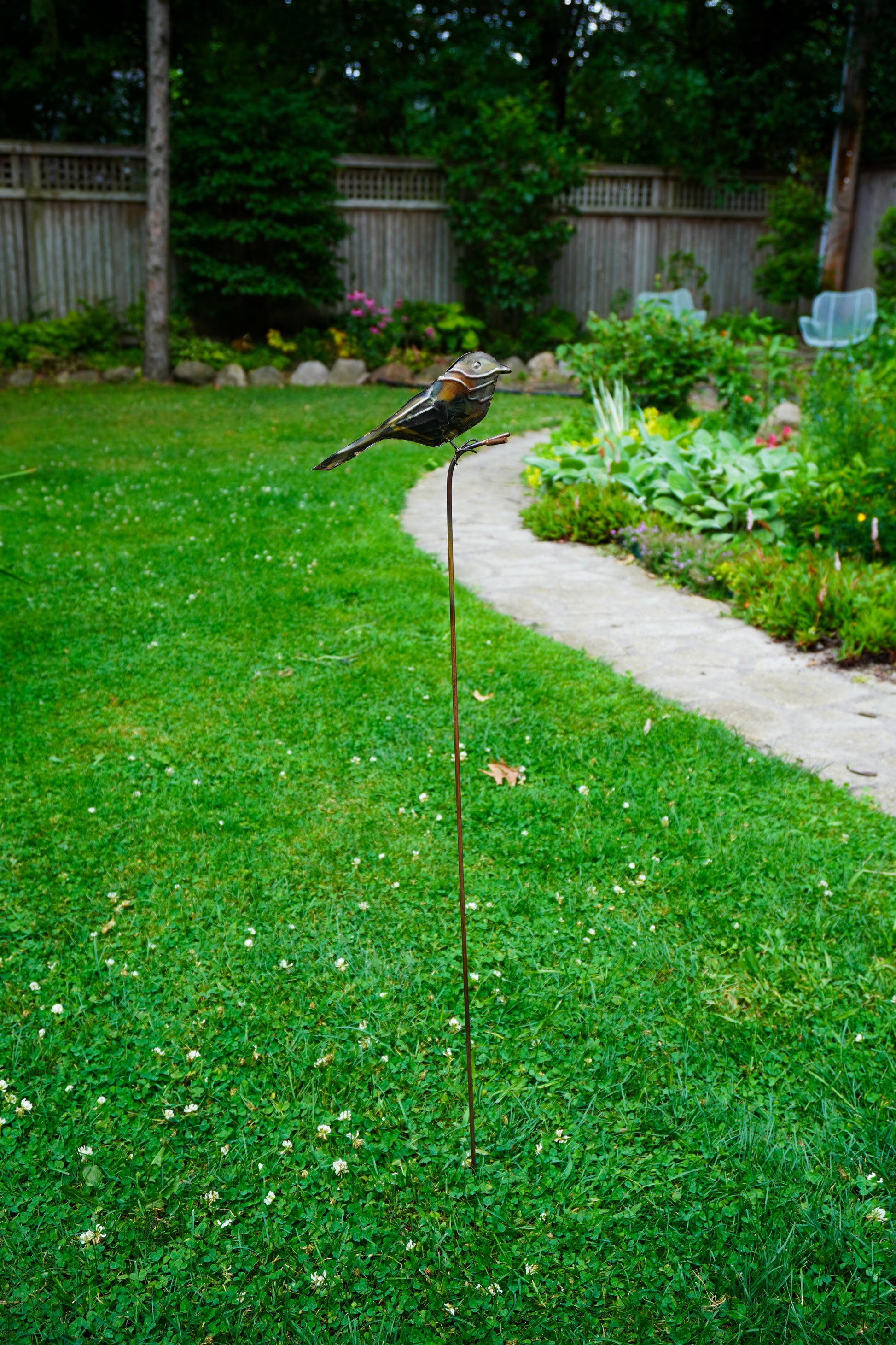 A metal garden bird statue perched on a thin stake in a green backyard lawn, with a garden path and fence in the background.