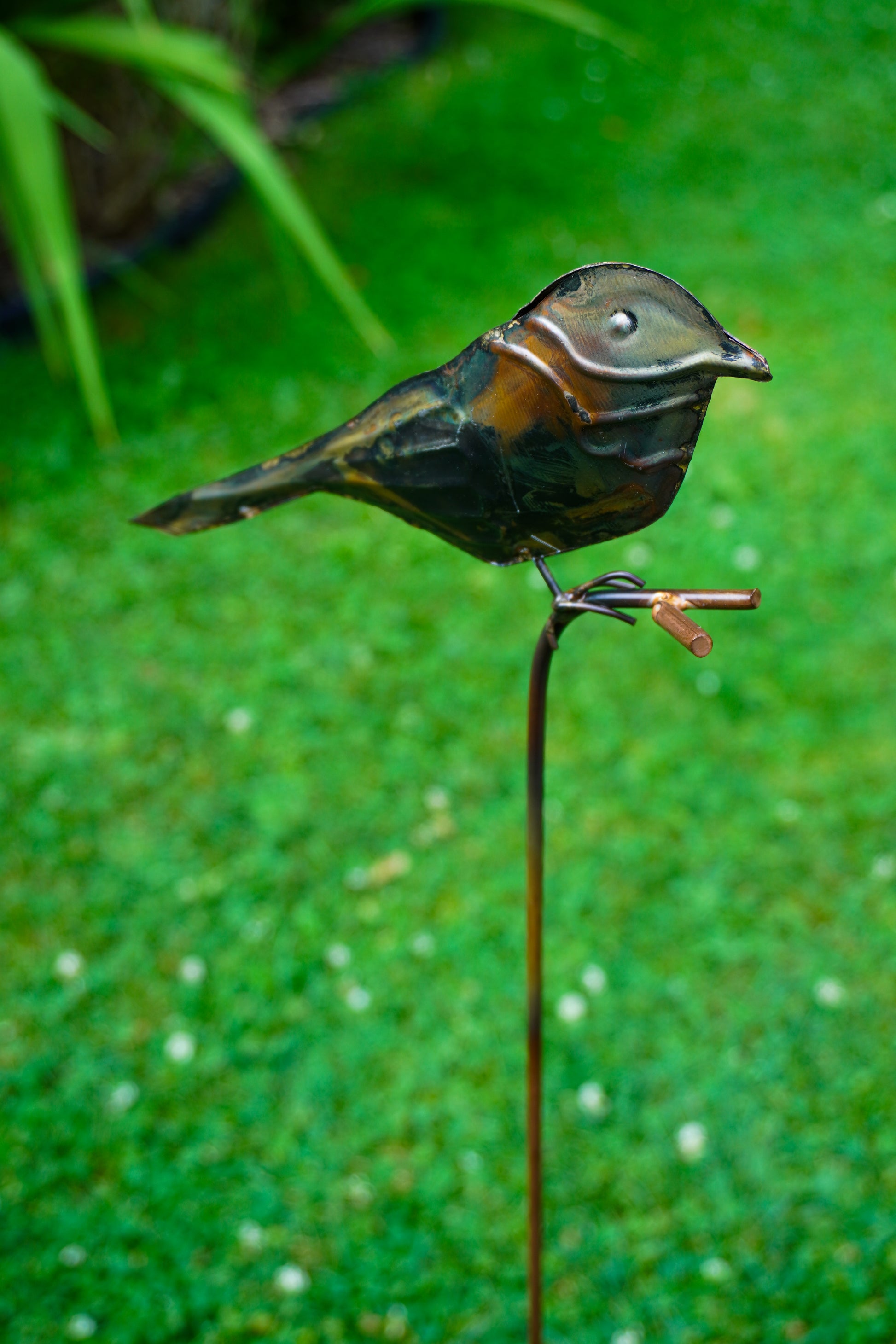 A metal bird garden stake sculpture perched on a thin stake, photographed outdoors against a blurred green grassy background.