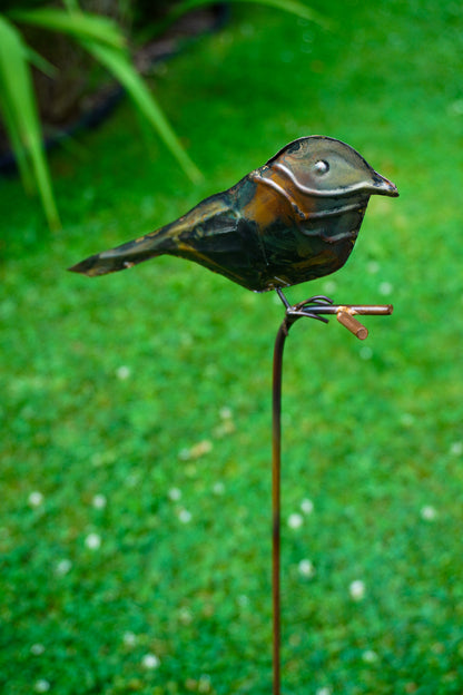 A metal bird garden stake sculpture perched on a thin stake, photographed outdoors against a blurred green grassy background.