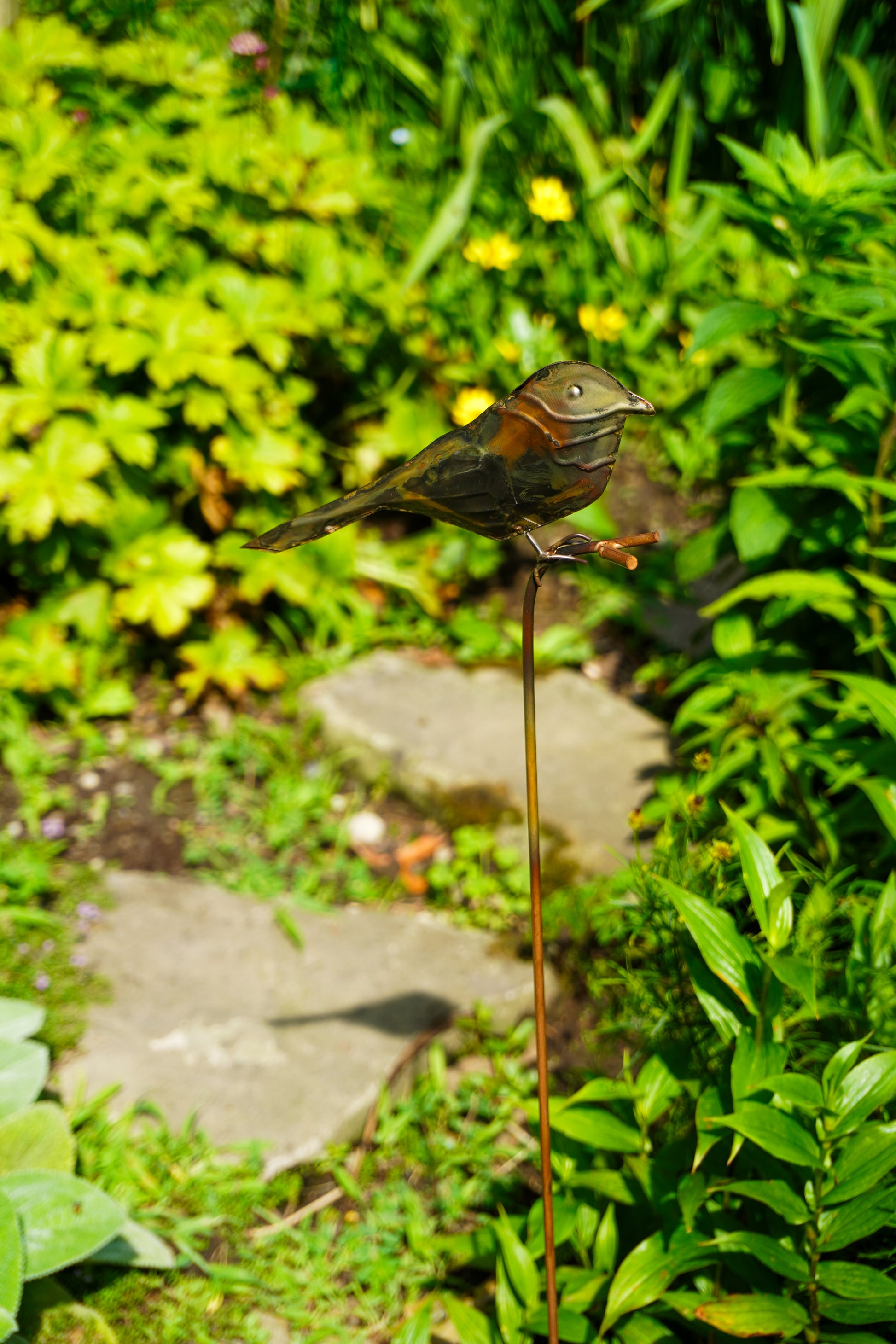 A small bird garden stake sculpture with a metallic bronze finish, perched on a thin stake in a lush green garden setting.