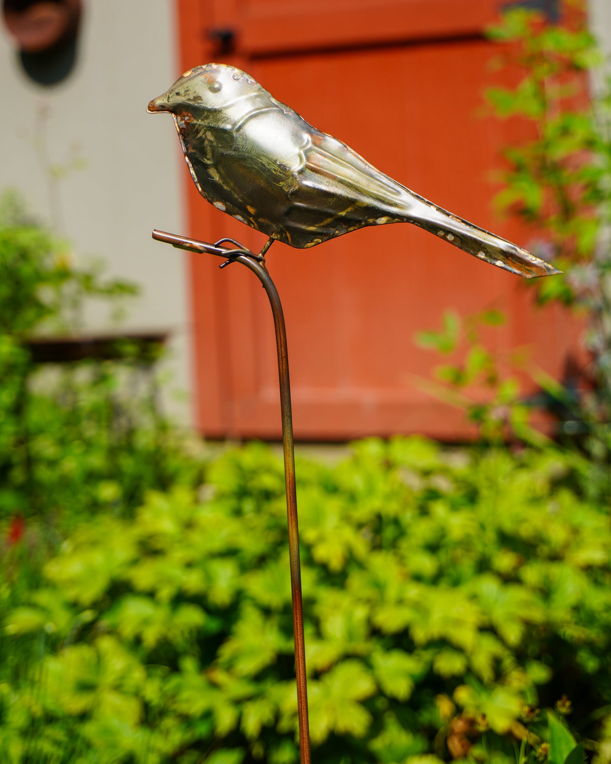 A metallic bird garden stake sculpture in a shiny silver finish, perched on a thin brown metal stake, set against a blurred background of green foliage and a red-orange door.