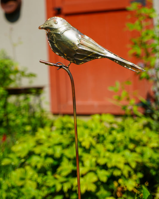 A metallic bird garden stake sculpture in a shiny silver finish, perched on a thin brown metal stake, set against a blurred background of green foliage and a red-orange door.
