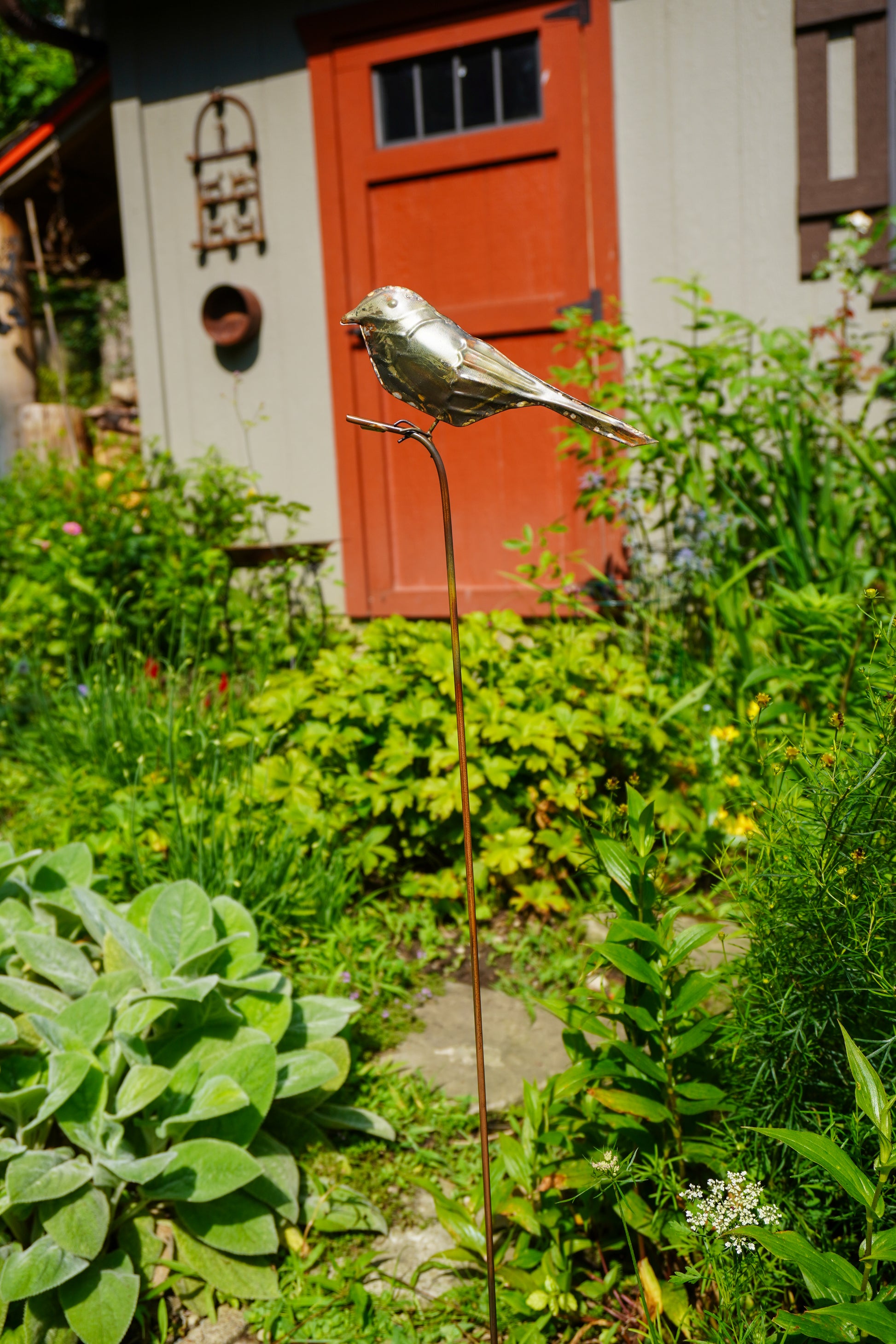 A metallic bird garden stake sculpture in a real garden setting with green plants, a stone path, and a red door in the background.