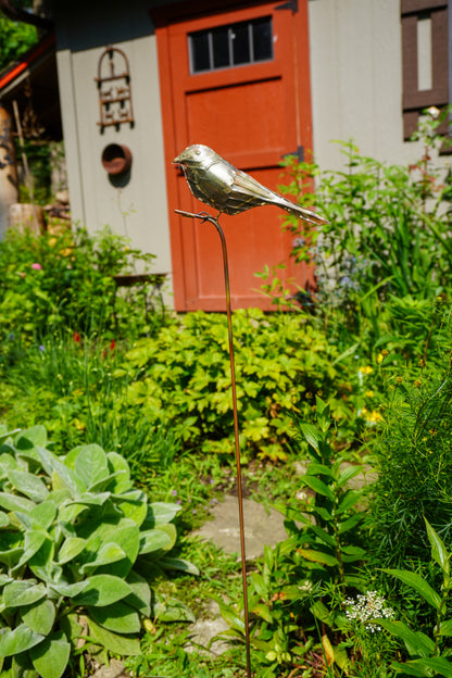 A metallic bird garden stake sculpture in a real garden setting with green plants, a stone path, and a red door in the background.