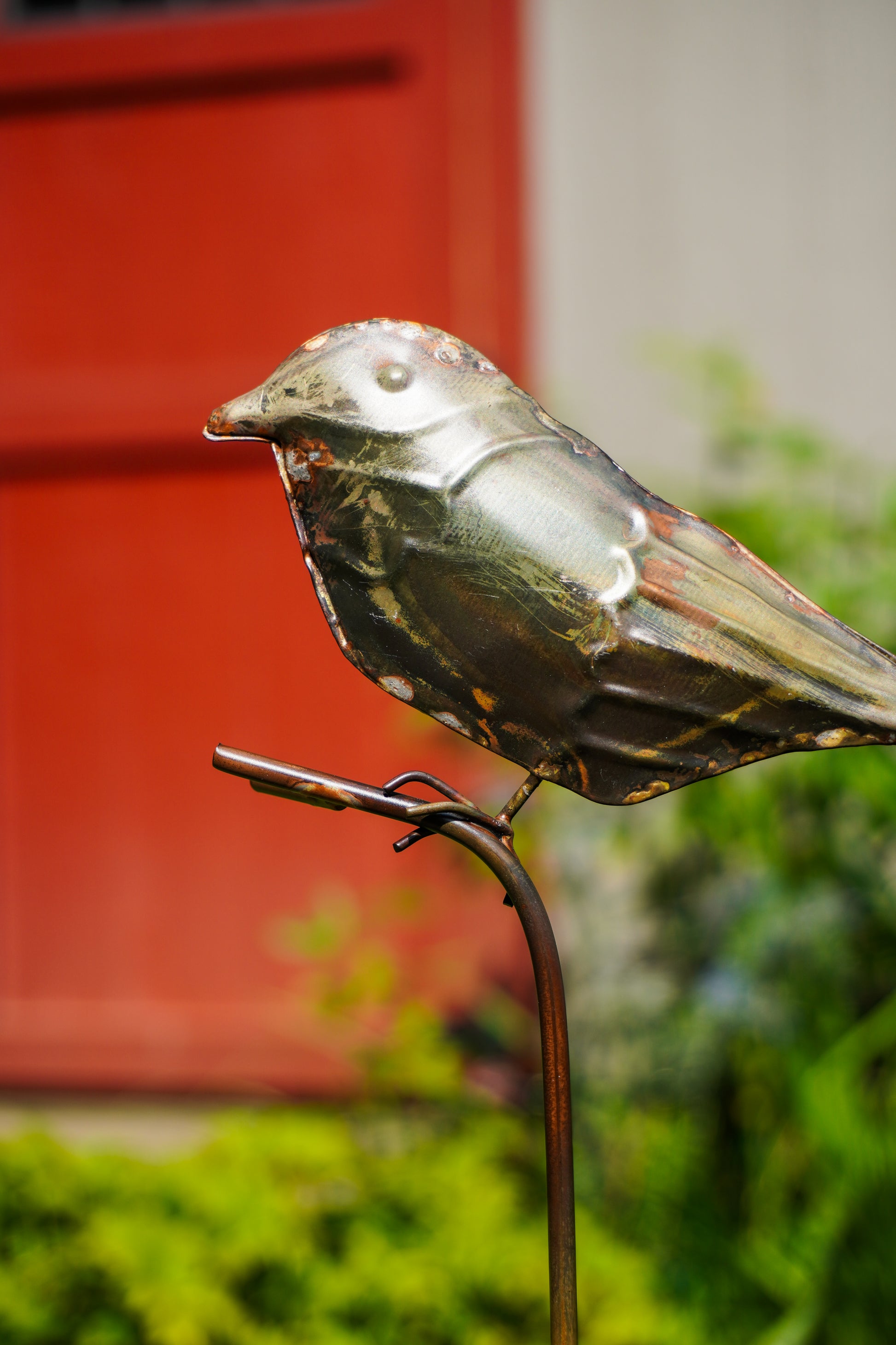 A metallic bird garden sculpture perched on a thin metal stake, photographed outdoors with a blurred red door and green foliage in the background.