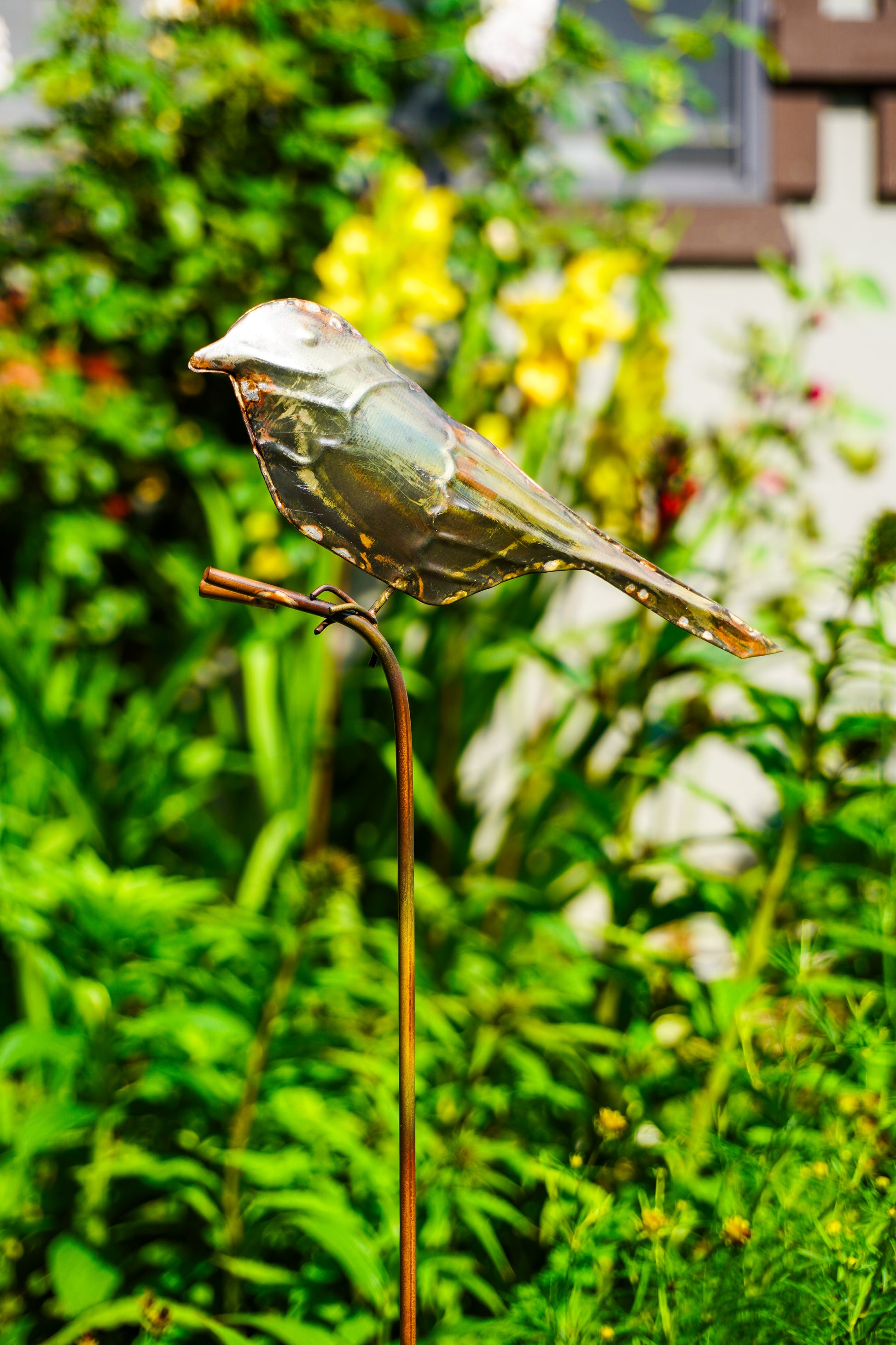 Metallic bird garden stake sculpture perched on a thin vertical stake, photographed outdoors in a lush green garden with blurred foliage and flowers in the background.