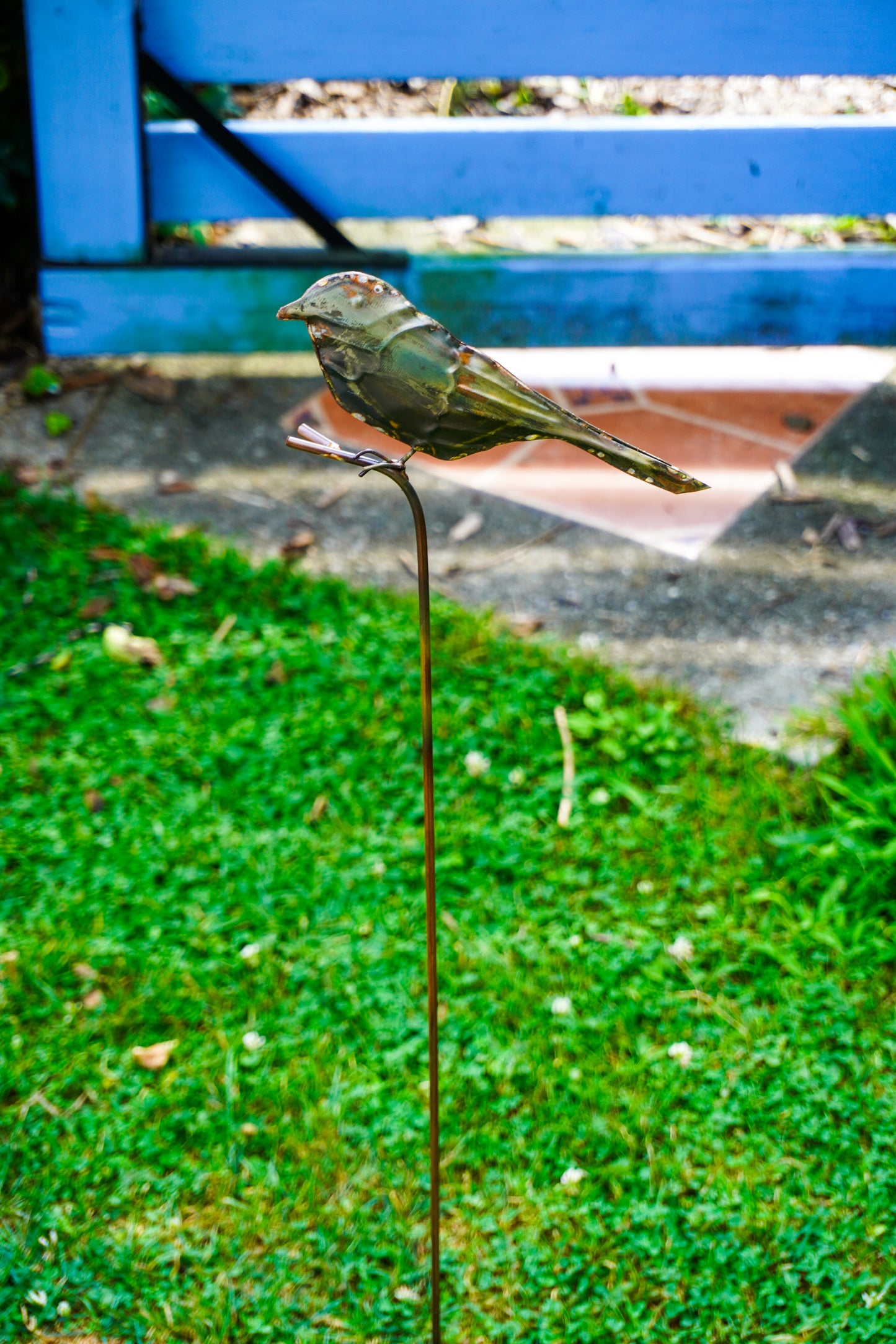 A metal bird garden stake sculpture of a small brown bird perched on a thin vertical stake, placed on green grass with a blurred blue fence and patio background.