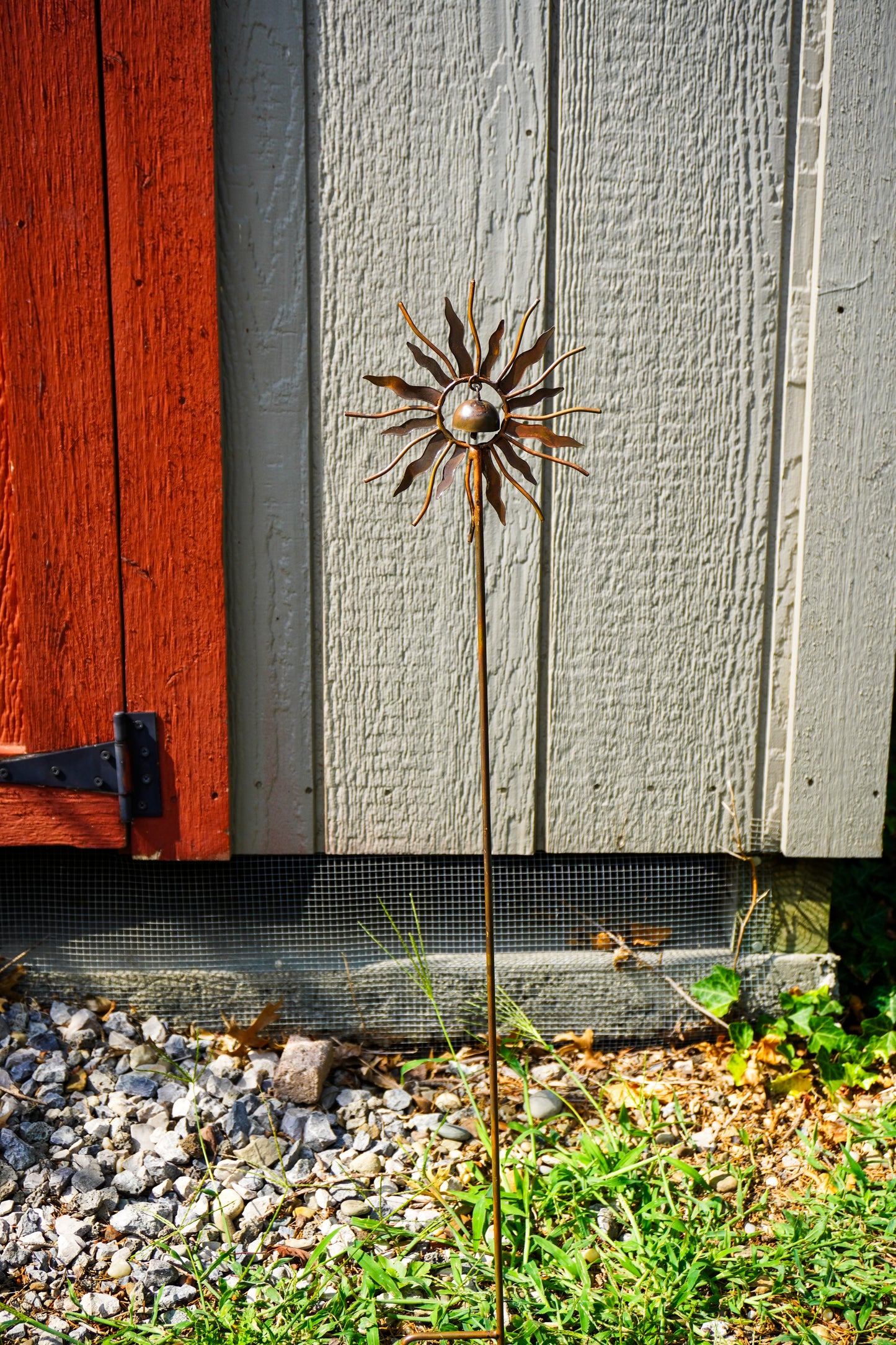 A metal sunflower garden stake sculpture with a textured bronze center and radiating sunburst petals, planted in a gravel and grassy area in front of a gray wooden fence and a red door.