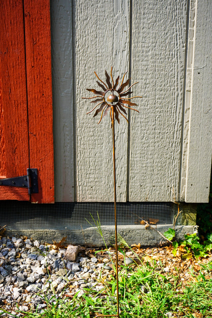 A metal sunflower garden stake sculpture with a textured bronze center and radiating sunburst petals, planted in a gravel and grassy area in front of a gray wooden fence and a red door.