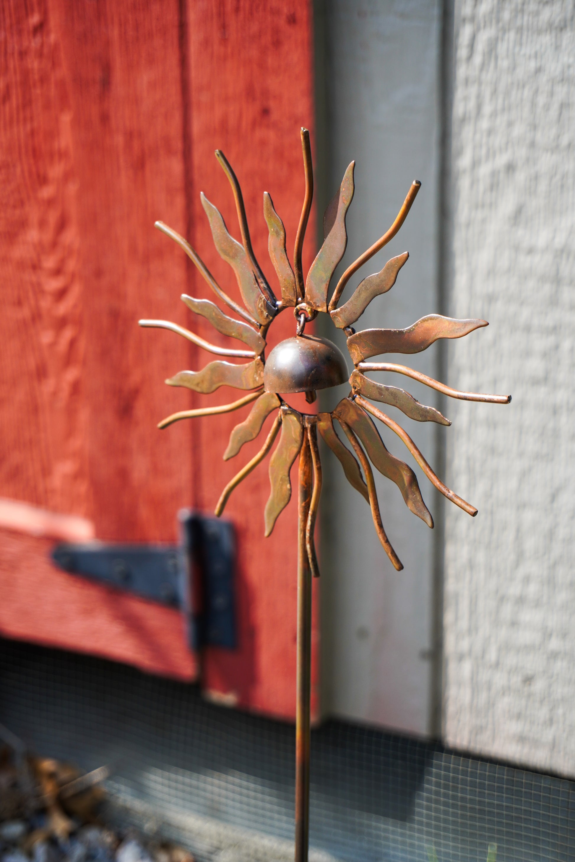 A metal sunflower garden stake sculpture with a polished silver center and rusted copper-toned petals, photographed outdoors in front of a red wooden door and light gray wall.