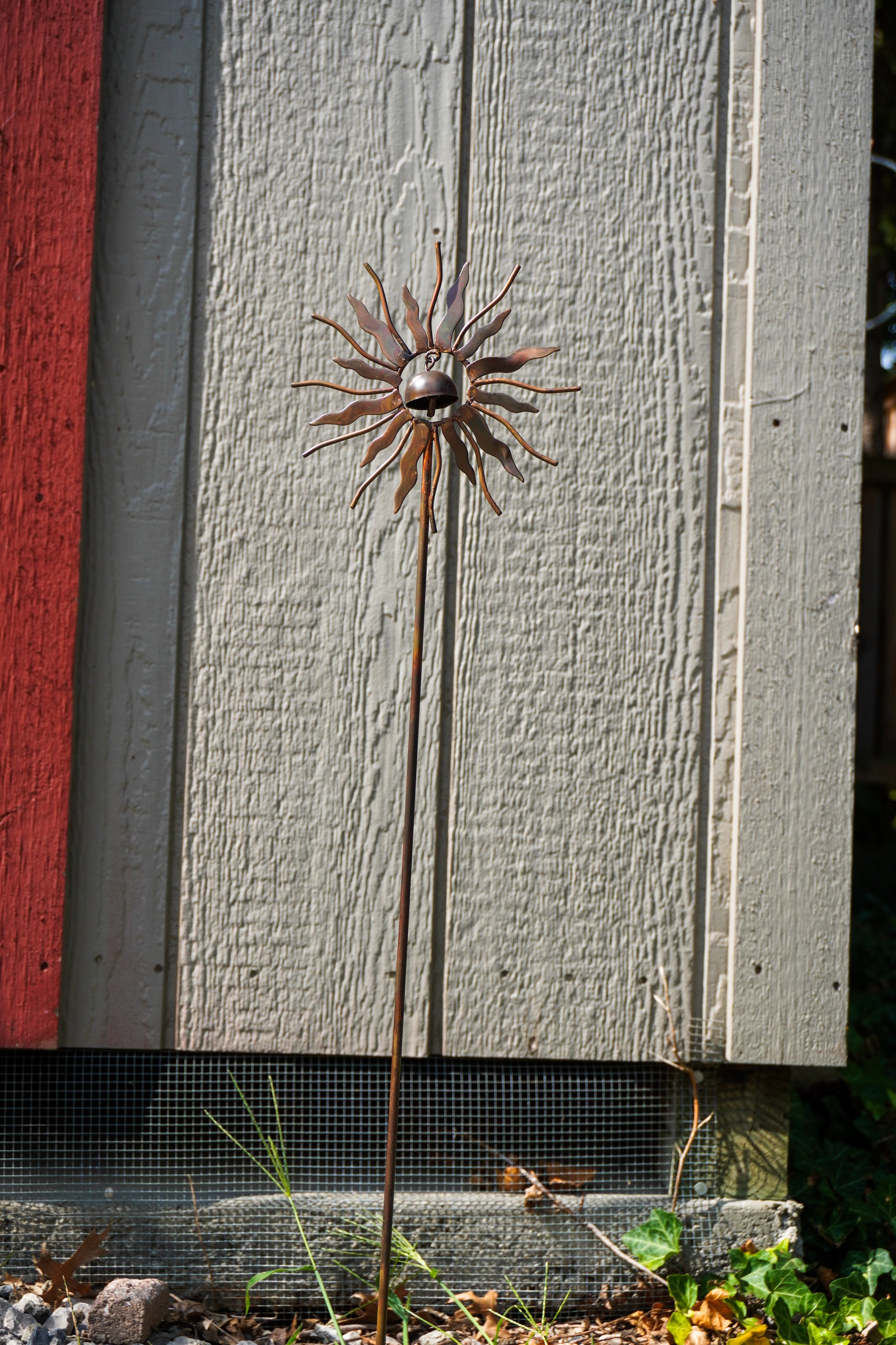 A metal sunflower garden stake sculpture with a rusted brown finish, standing outdoors in front of a gray wooden shed wall.