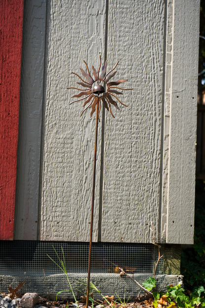 A metal sunflower garden stake sculpture with a rusted brown finish, standing outdoors in front of a gray wooden shed wall.
