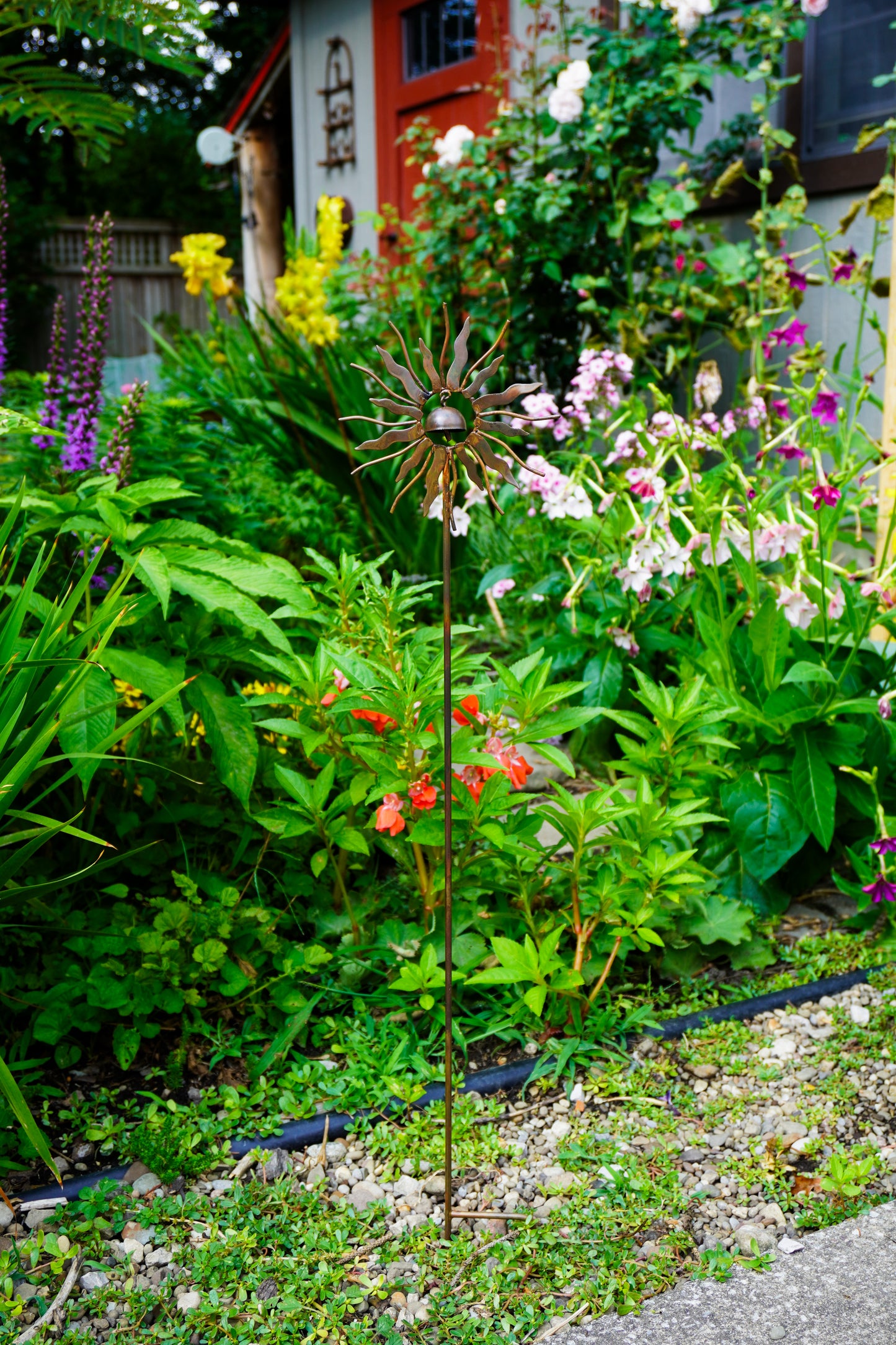 A metal sunflower garden stake with a decorative starburst flower in the center, planted in a lush green garden bed with other flowers and foliage.