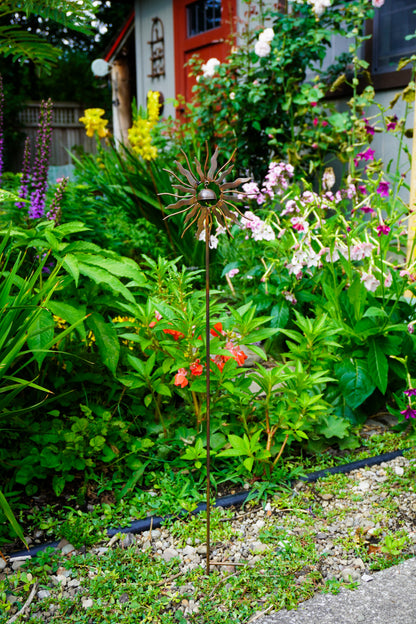A metal sunflower garden stake with a decorative starburst flower in the center, planted in a lush green garden bed with other flowers and foliage.
