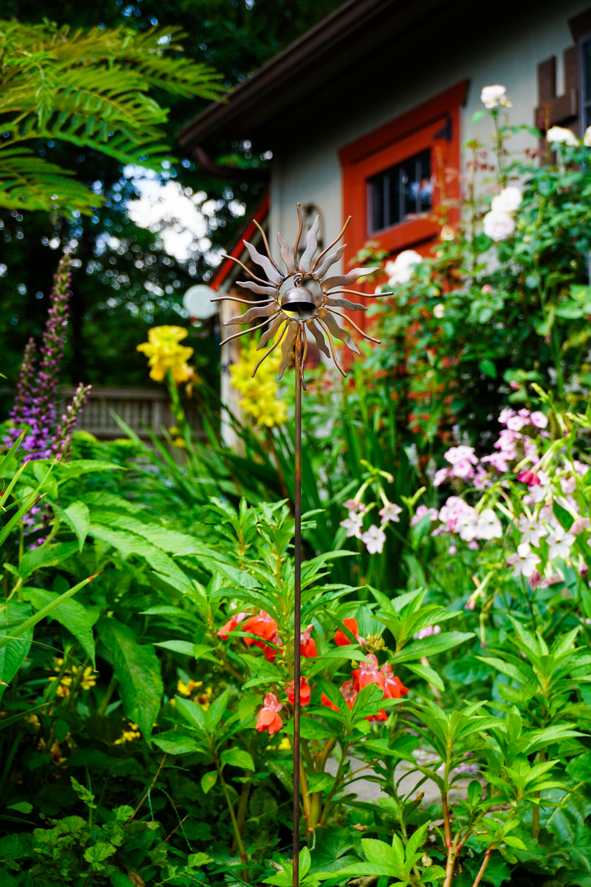 A metal sunburst garden stake sculpture with a bell at its center, displayed in a lush green garden with red, yellow, and pink flowers in the foreground and a house in the background.