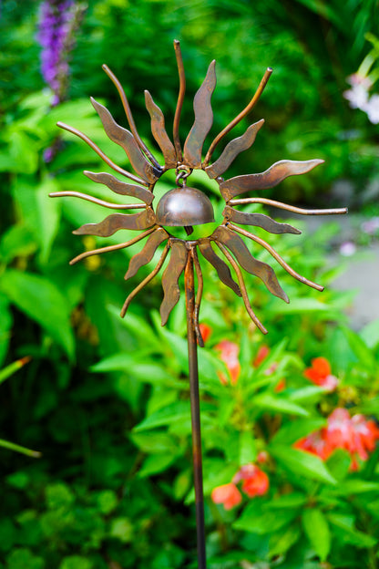 A metal sunflower garden stake sculpture with rusted brown petals and a glossy green glass center, photographed outdoors against a blurred green garden background with small red flowers.