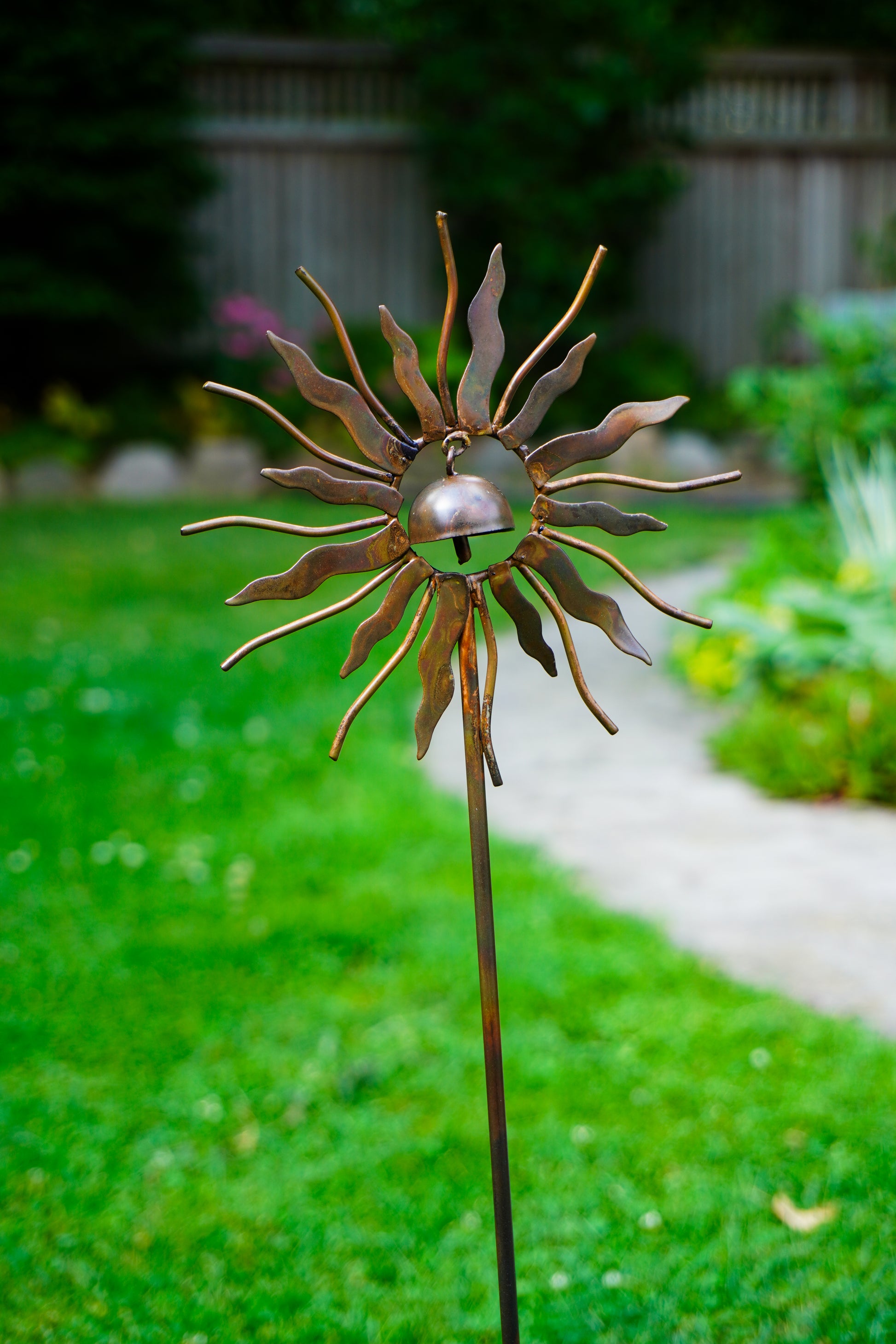 A metal sunflower garden stake sculpture with a small bell inside the center, set in a green backyard garden.