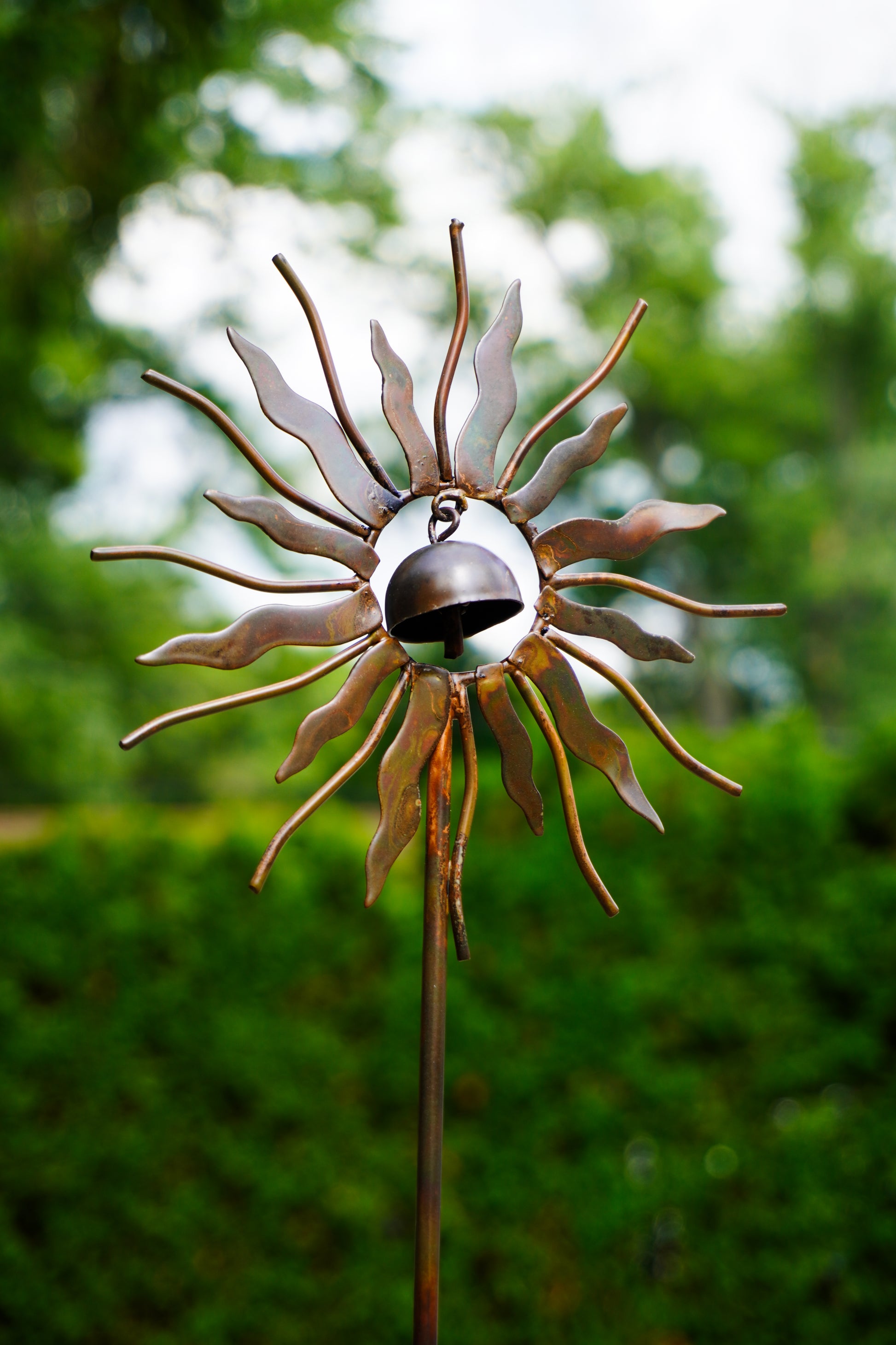A metal sunflower garden stake ornament with a small hanging bell in the center, photographed outdoors against a blurred green garden background.