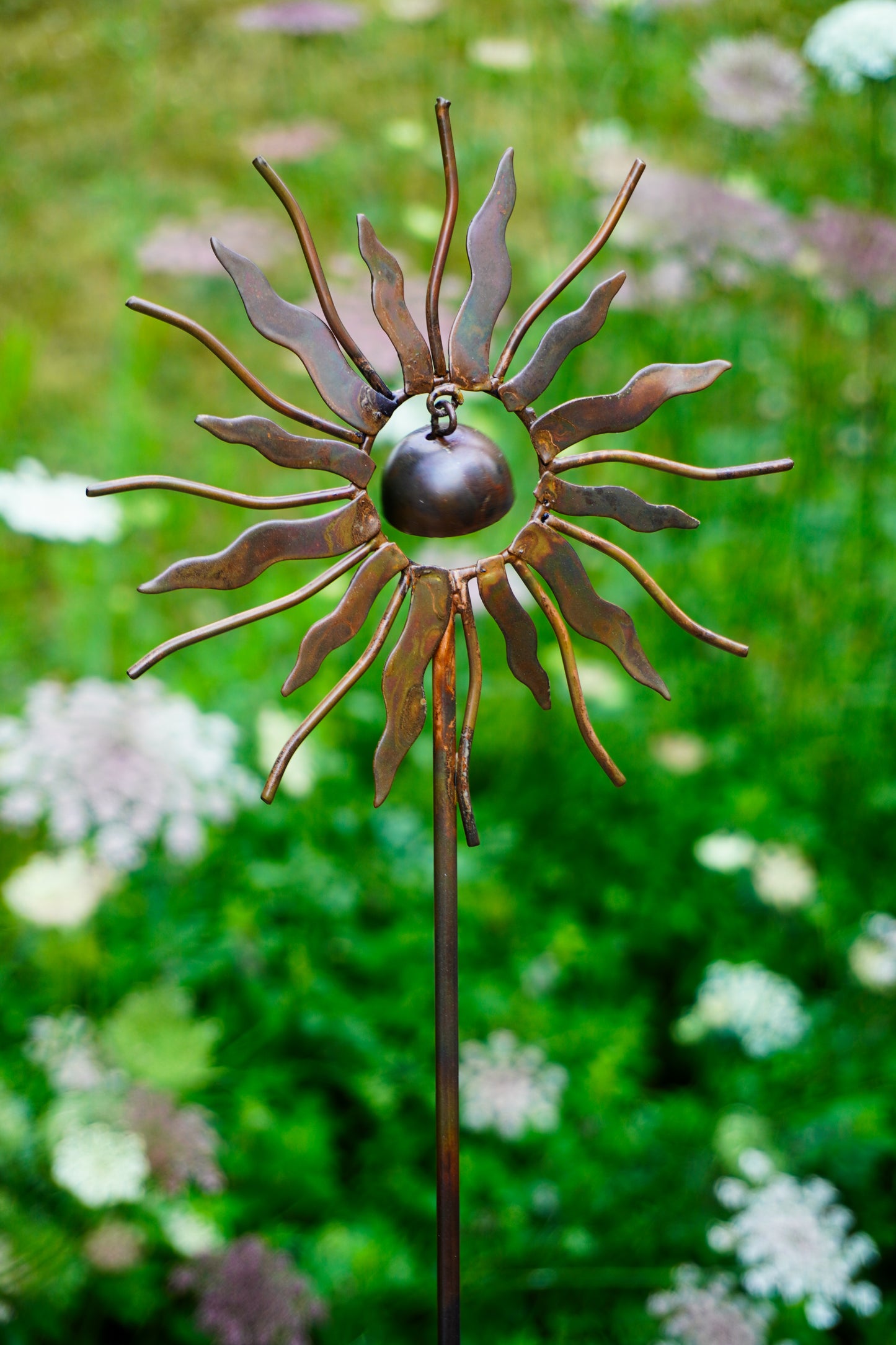 A metal sunflower garden stake sculpture with a dark bronze center and rusted brown petals, set against a blurred green garden background with white flowers.