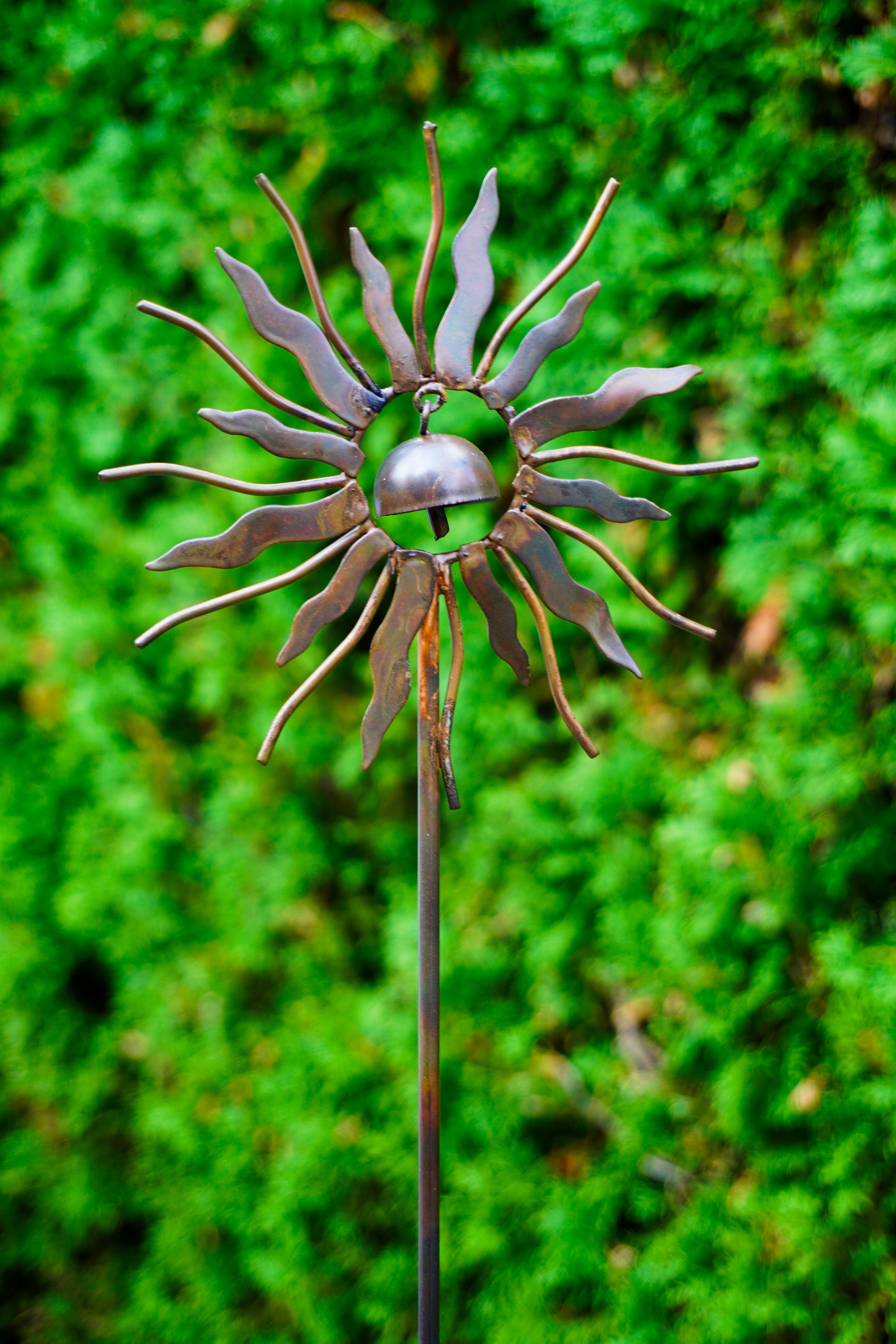 A metal sunflower garden stake sculpture with a small hanging bell in the center, photographed outdoors against a blurred green foliage background.