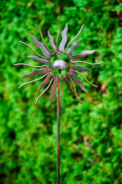 A metal sunflower garden stake sculpture with a small hanging bell in the center, photographed outdoors against a blurred green foliage background.
