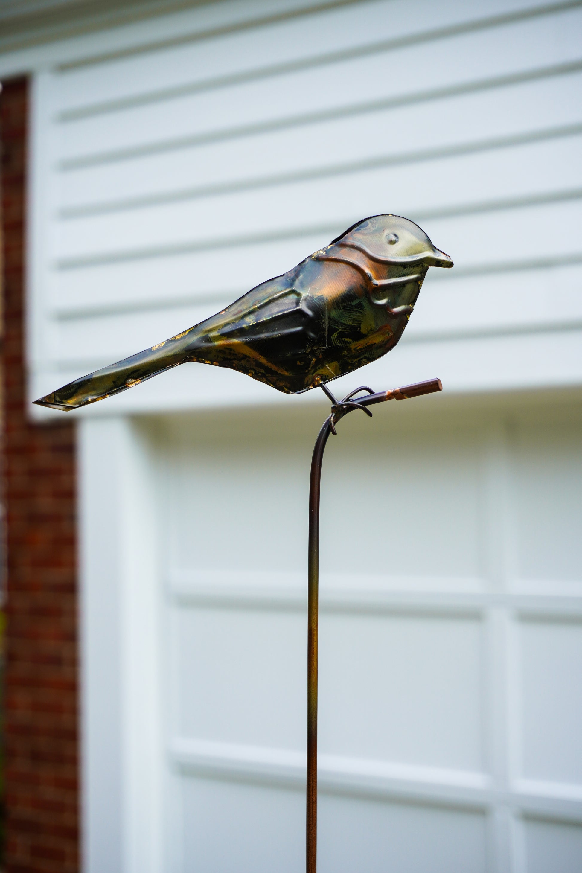 Metallic bird garden stake sculpture perched on a thin vertical stake, photographed outdoors in front of a white garage door and brick wall.
