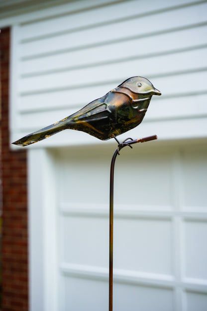 Metallic bird garden stake sculpture perched on a thin vertical stake, photographed outdoors in front of a white garage door and brick wall.