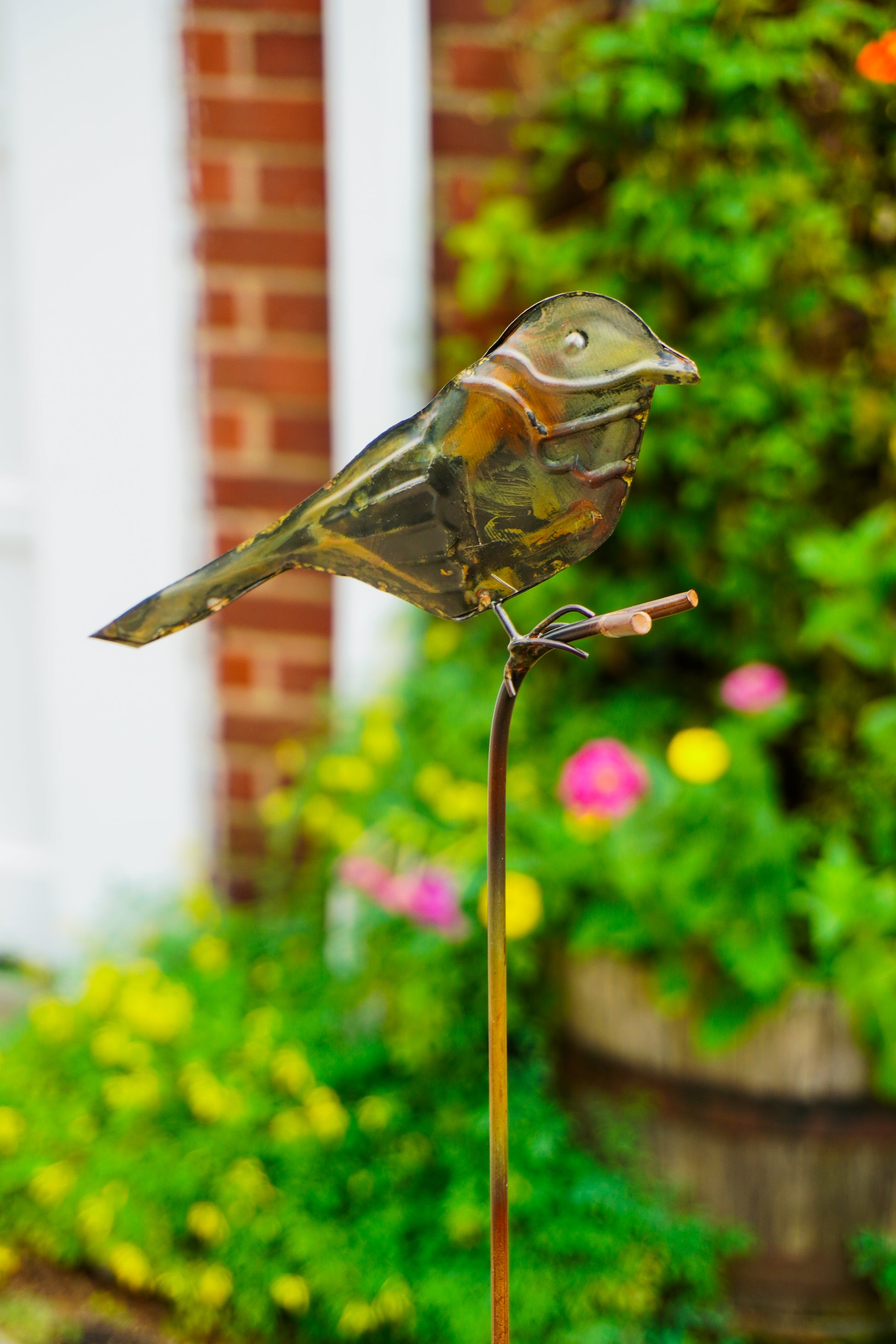 A metal bird garden stake sculpture with a distressed bronze finish, perched on a thin metal stake in a blurred garden setting.