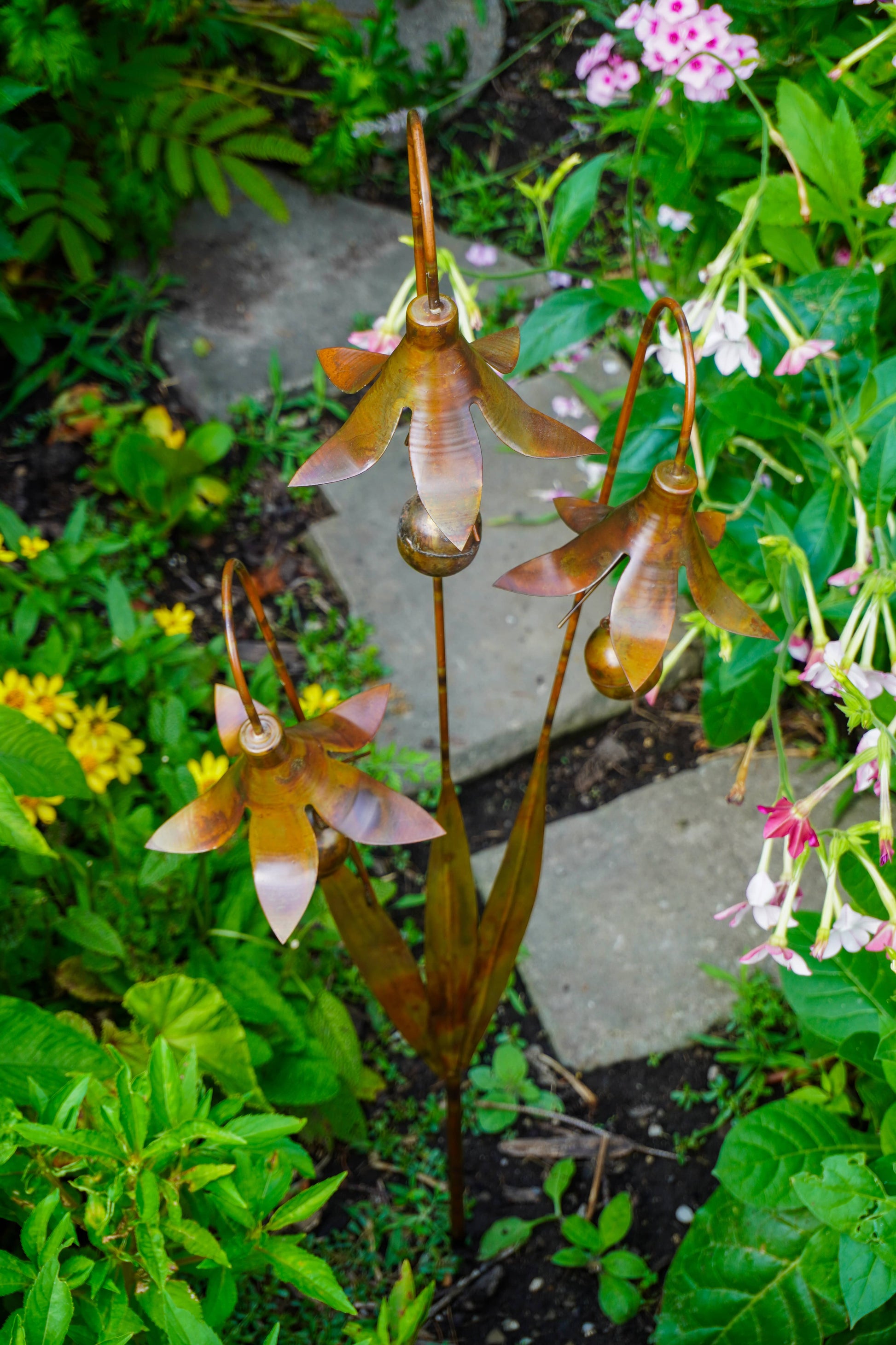 Three metal garden sculptures shaped like stylized flowers on long stems, displayed in a lush green garden next to a stone path.