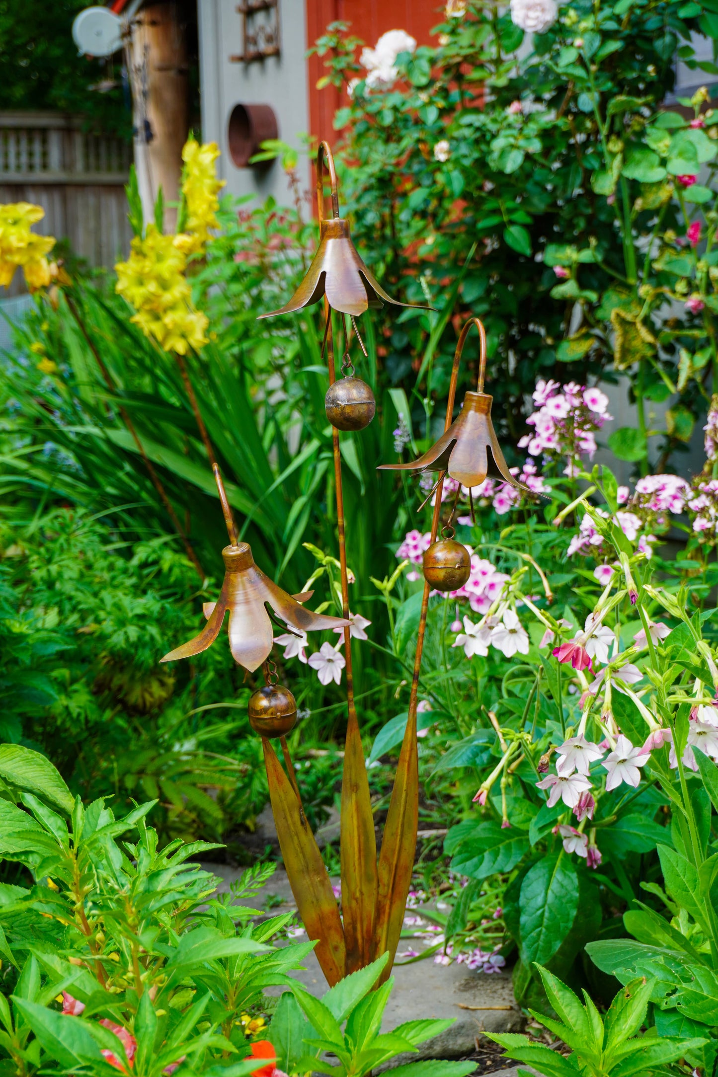 A garden stake sculpture featuring bronze-toned metal flower heads with hanging bell-like elements and brown leaf-like bases, set in a lush garden with green foliage, pink flowers, and yellow blooms.