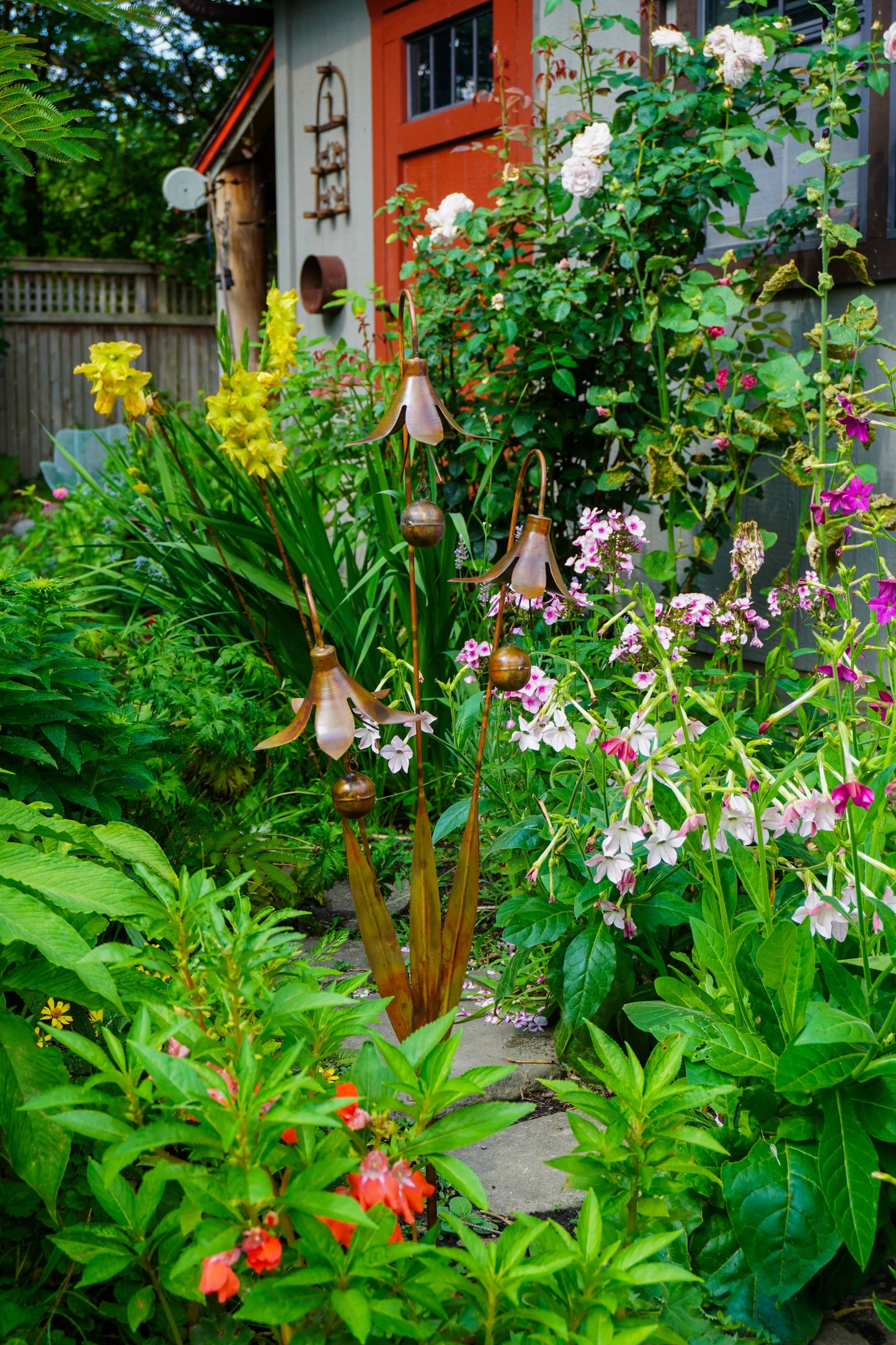 A lush garden scene with dense green foliage, pink and purple flowers, yellow flowers, and a small metal garden sculpture in the center. A house exterior with a red door is visible in the background.