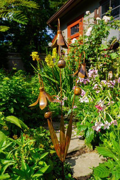 A bronze metal garden sculpture of stylized flowers and leaves, set in a lush green garden with a stone path and house in the background.
