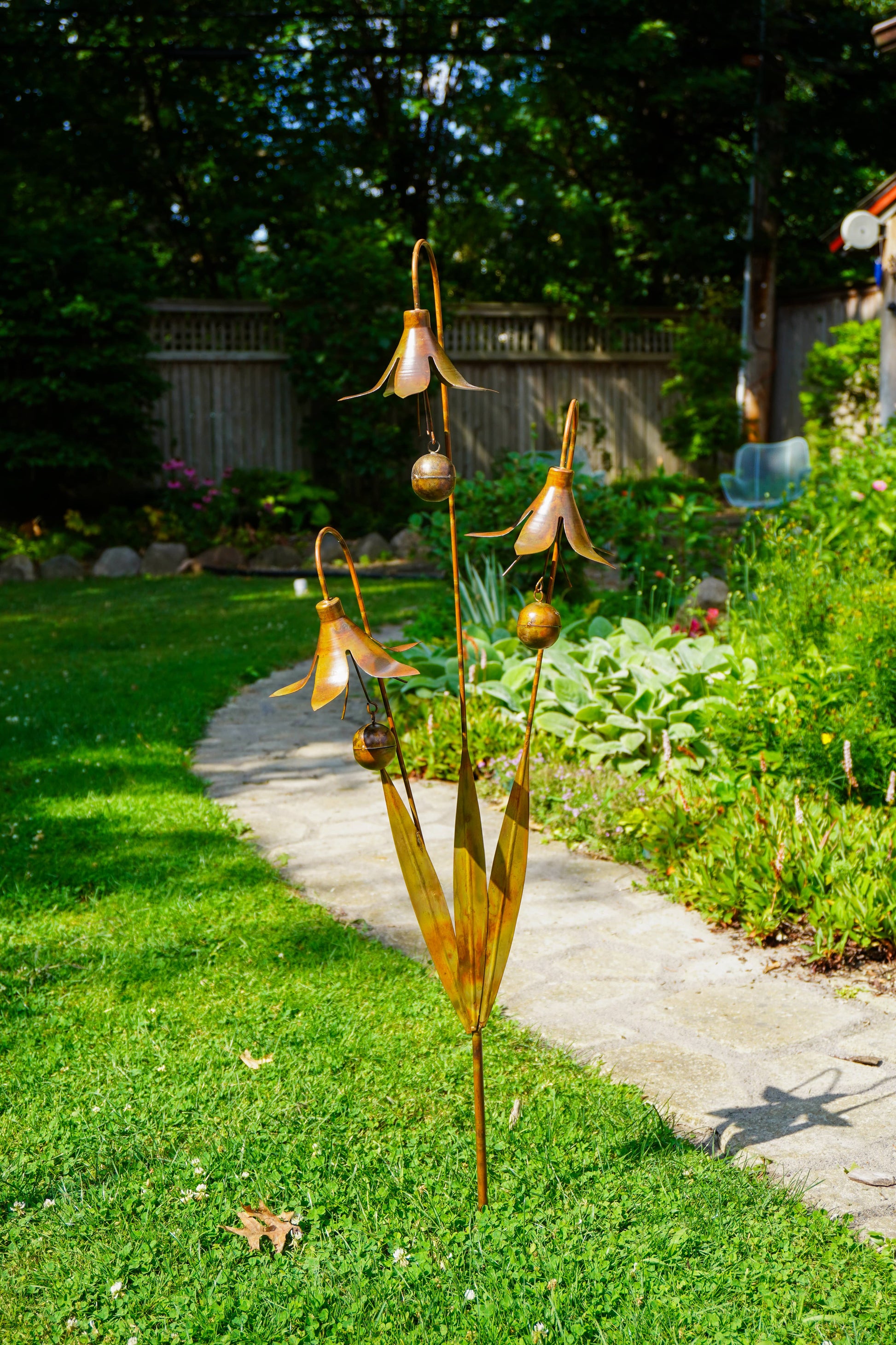 A metal garden sculpture shaped like dried corn husks with hanging bell-shaped flowers and small round pods, displayed on a grassy lawn next to a concrete path in a backyard garden.