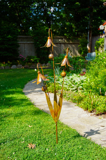 A metal garden sculpture shaped like dried corn husks with hanging bell-shaped flowers and small round pods, displayed on a grassy lawn next to a concrete path in a backyard garden.