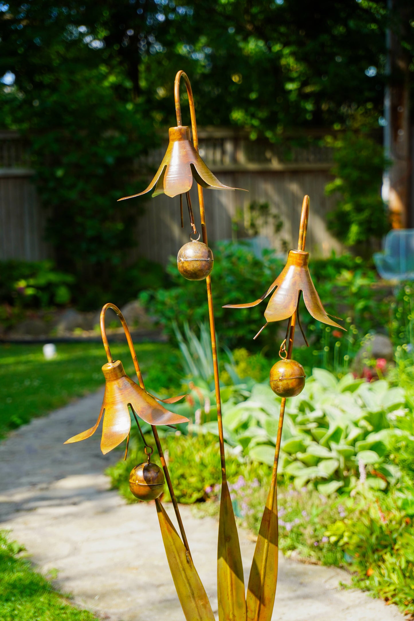 Metallic garden sculpture of three bronze bell-shaped flowers with hanging bells, set in a lush green backyard garden on a stone path.